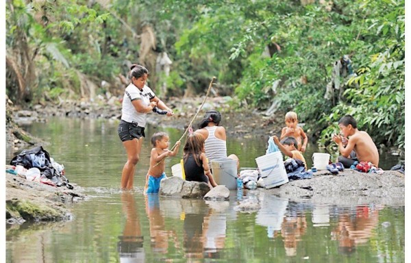 Mujeres lavan mientras niños se bañan en la quebrada.